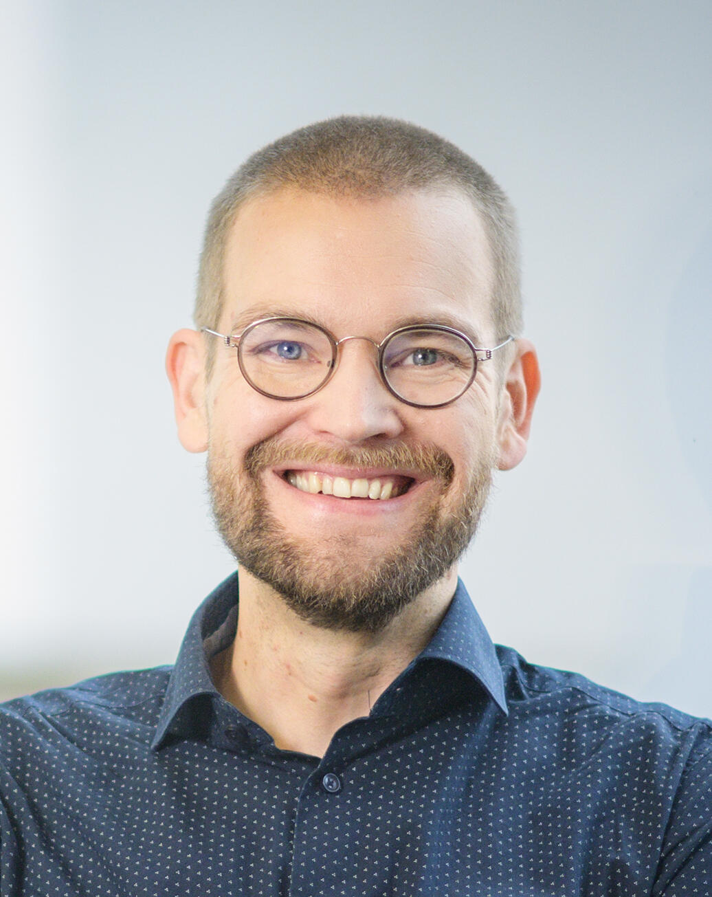 Smiling man with glasses, a beard, and short hair. He is wearing a dark blue button-up shirt, with a light-colored wall in the background.